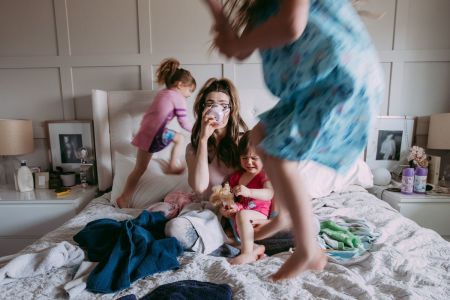 Photo of stressed out young mother sipping on a cup of coffee on her messy bed while her three daughters are jumping around her.